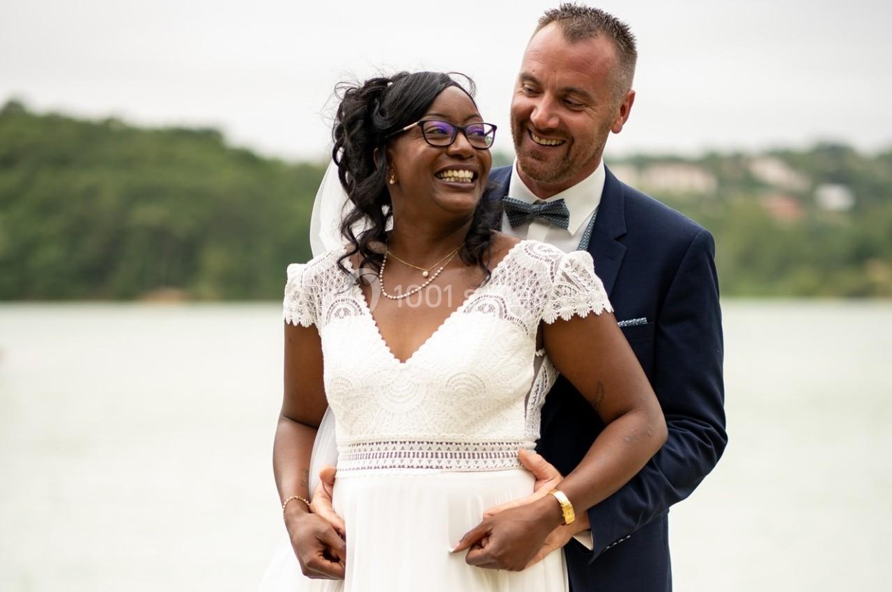 Un couple en tenue de mariage sourit devant un paysage naturel avec un lac et des arbres en arrière-plan.