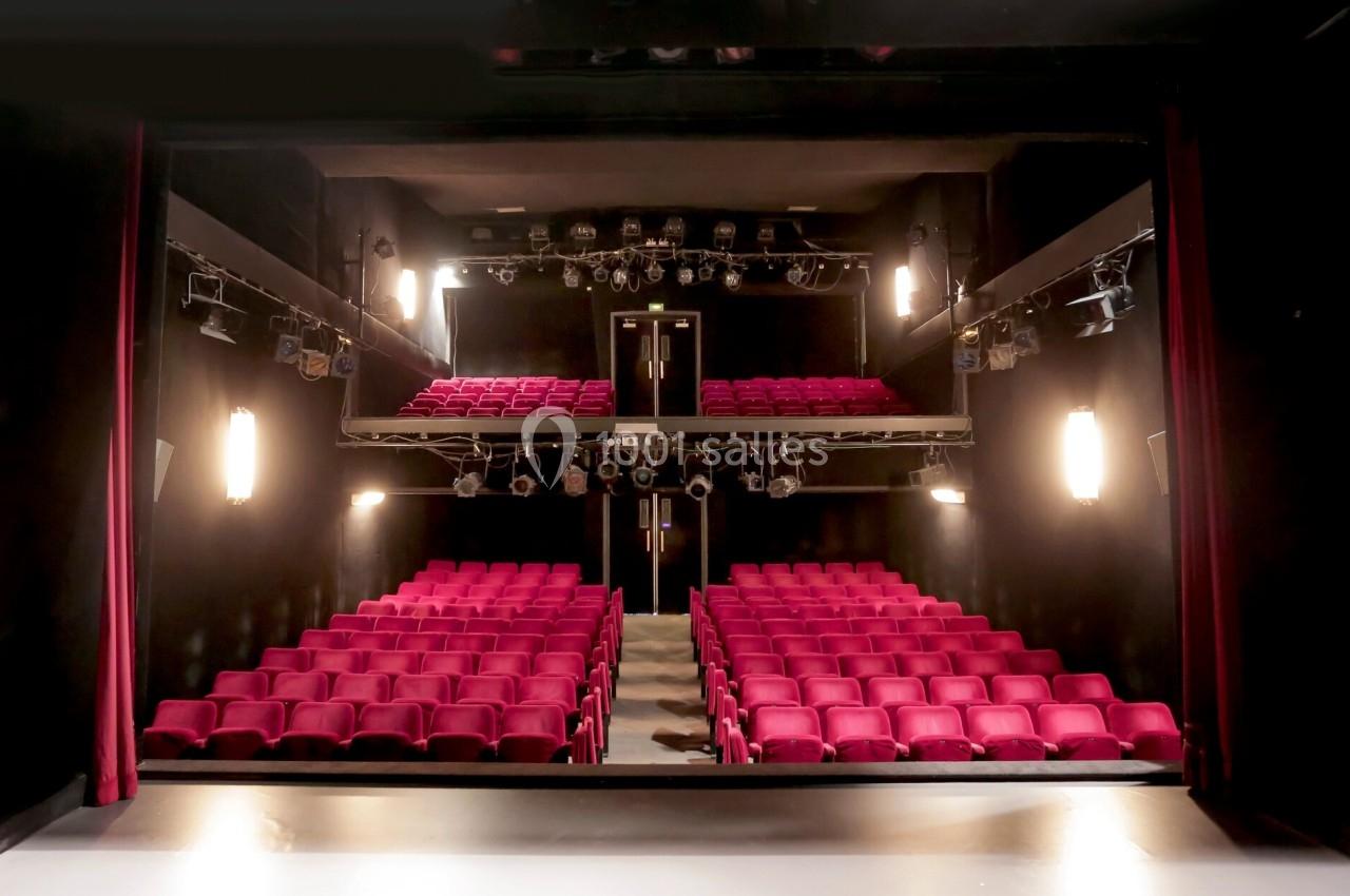 Salle de théâtre avec rangées de sièges rouges, scène au premier plan et éclairages suspendus.