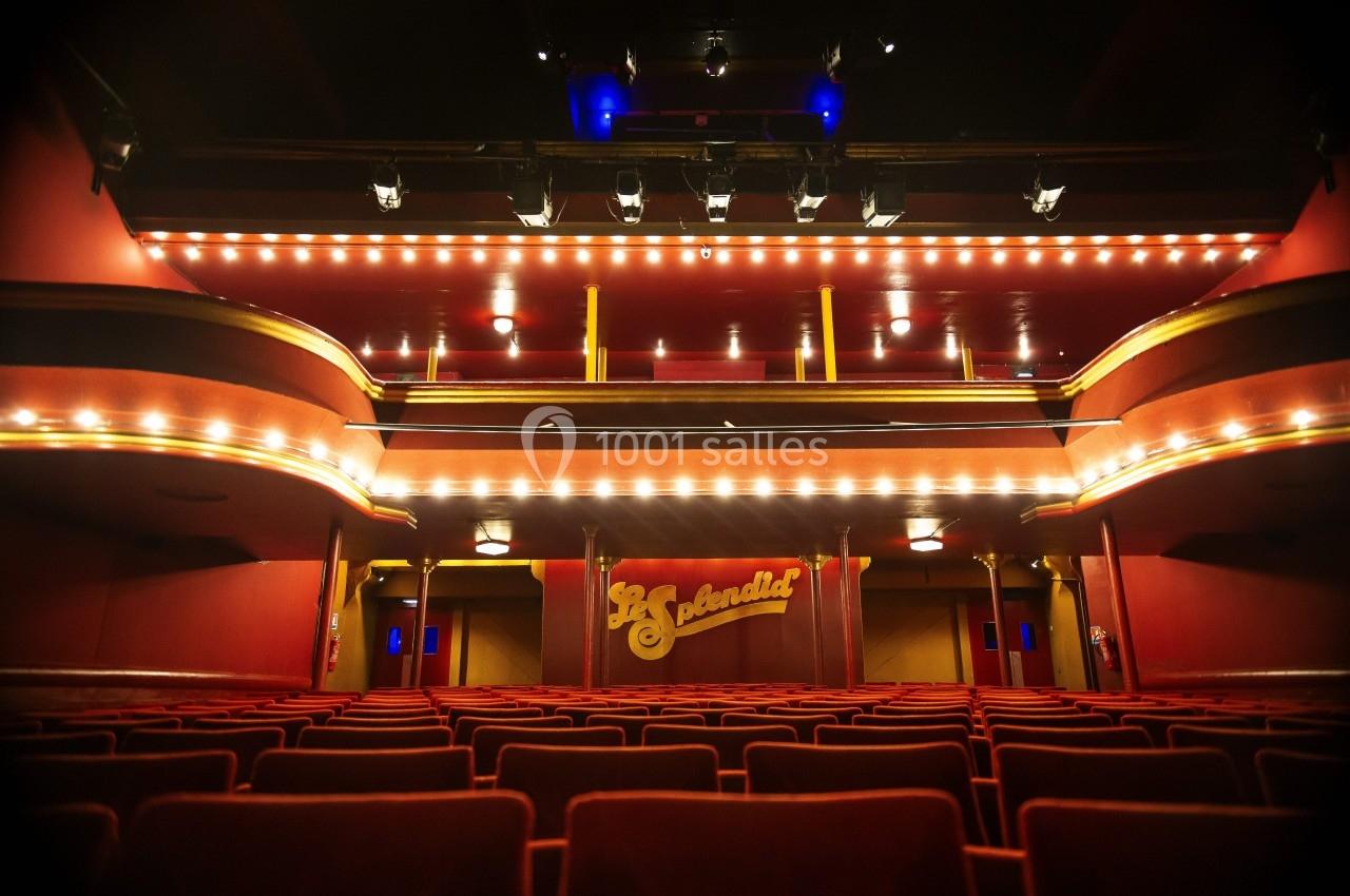 Salle de théâtre vide avec des rangées de sièges rouges, balcons éclairés et une scène ornée d'un rideau rouge.
