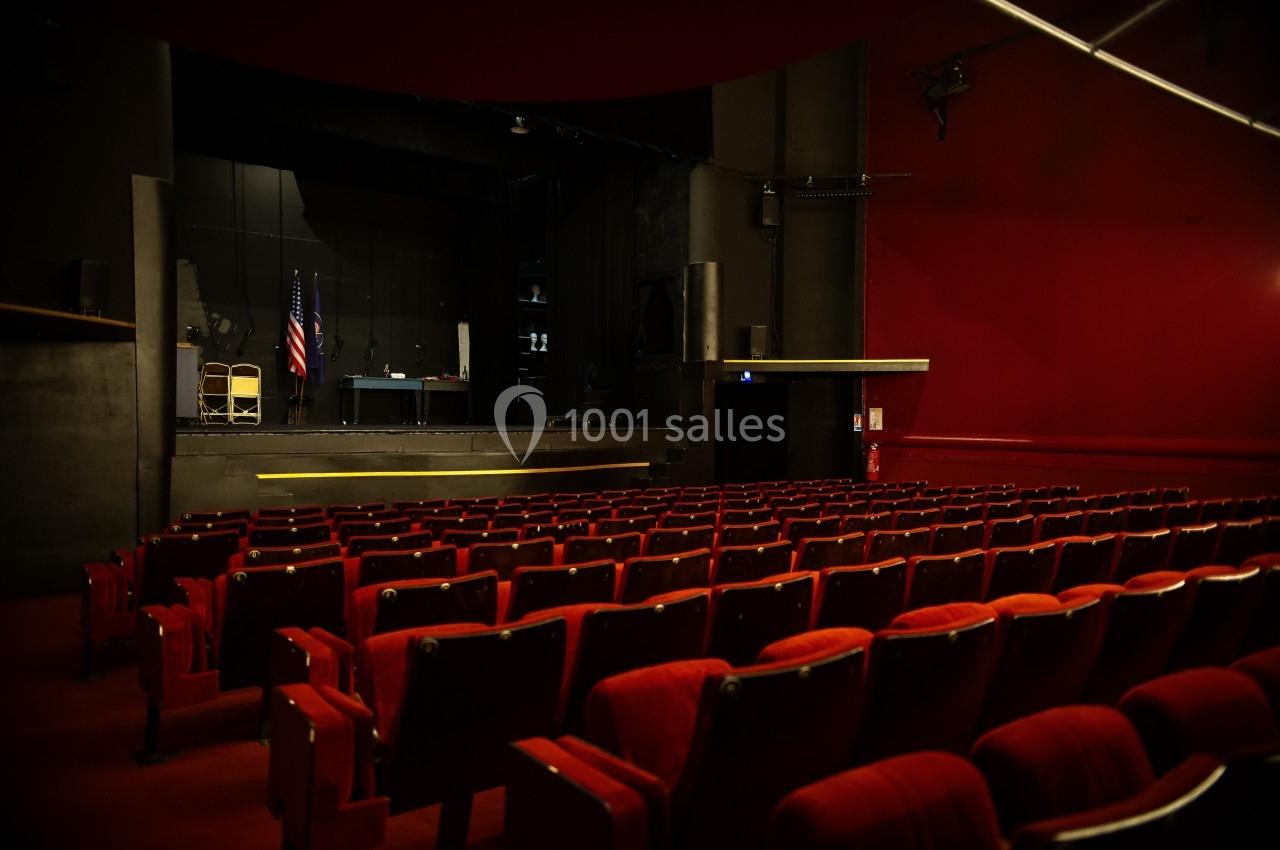 Salle de théâtre vide avec des rangées de sièges rouges face à une scène sombre équipée de quelques accessoires.