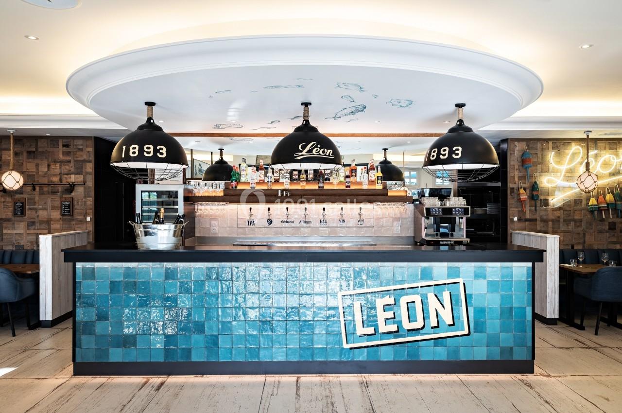 Comptoir de bar avec carrelage bleu, enseigne ’LEON’, luminaires suspendus et étagères avec bouteilles à l'arrière.