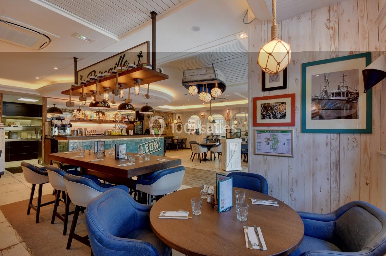 Salle de restaurant lumineuse avec tables dressées, chaises colorées et décoration murale maritime.