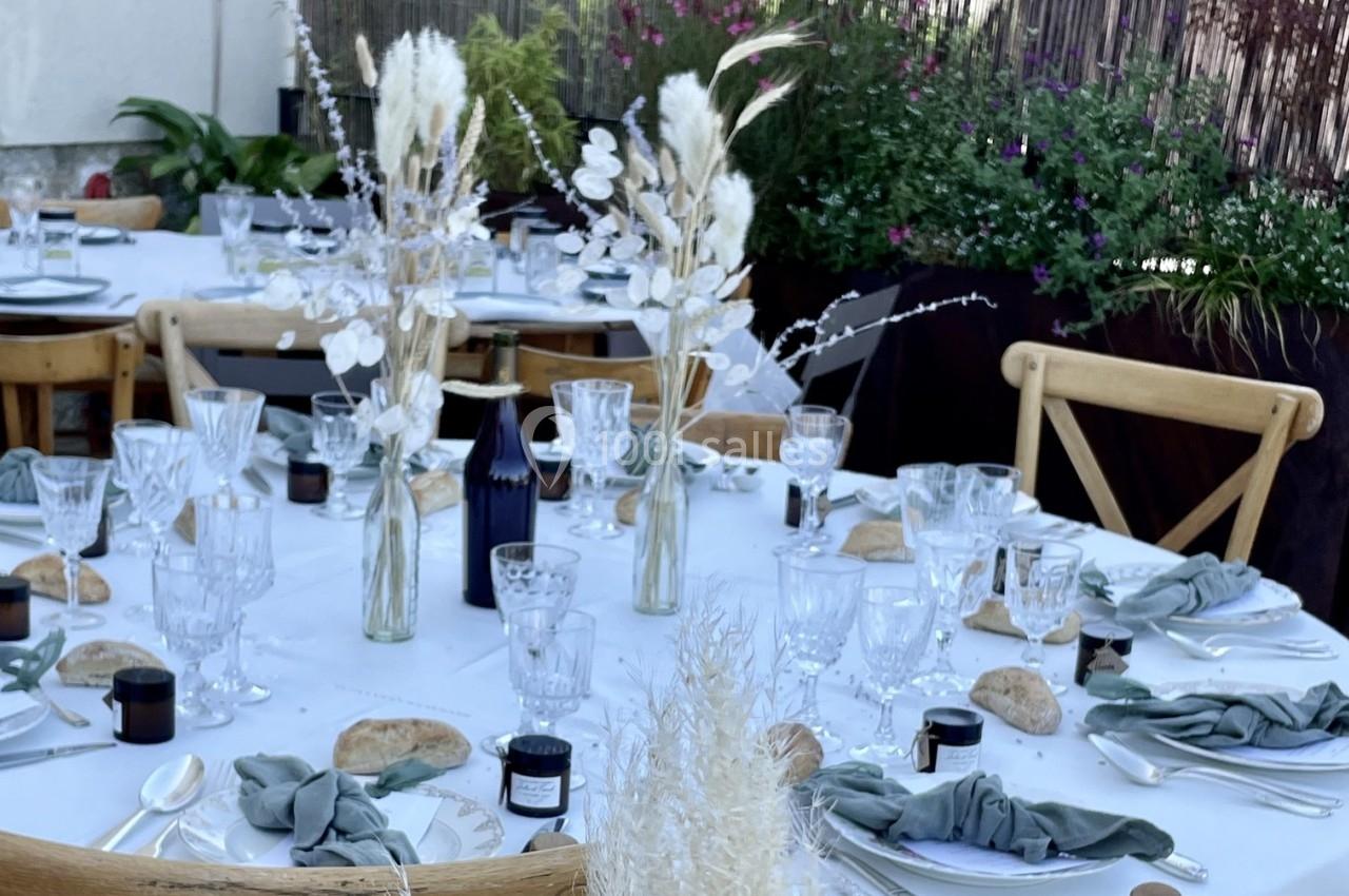 Table dressée en extérieur avec nappes blanches, verres, couverts, bouquets de fleurs séchées et décorations naturelles.