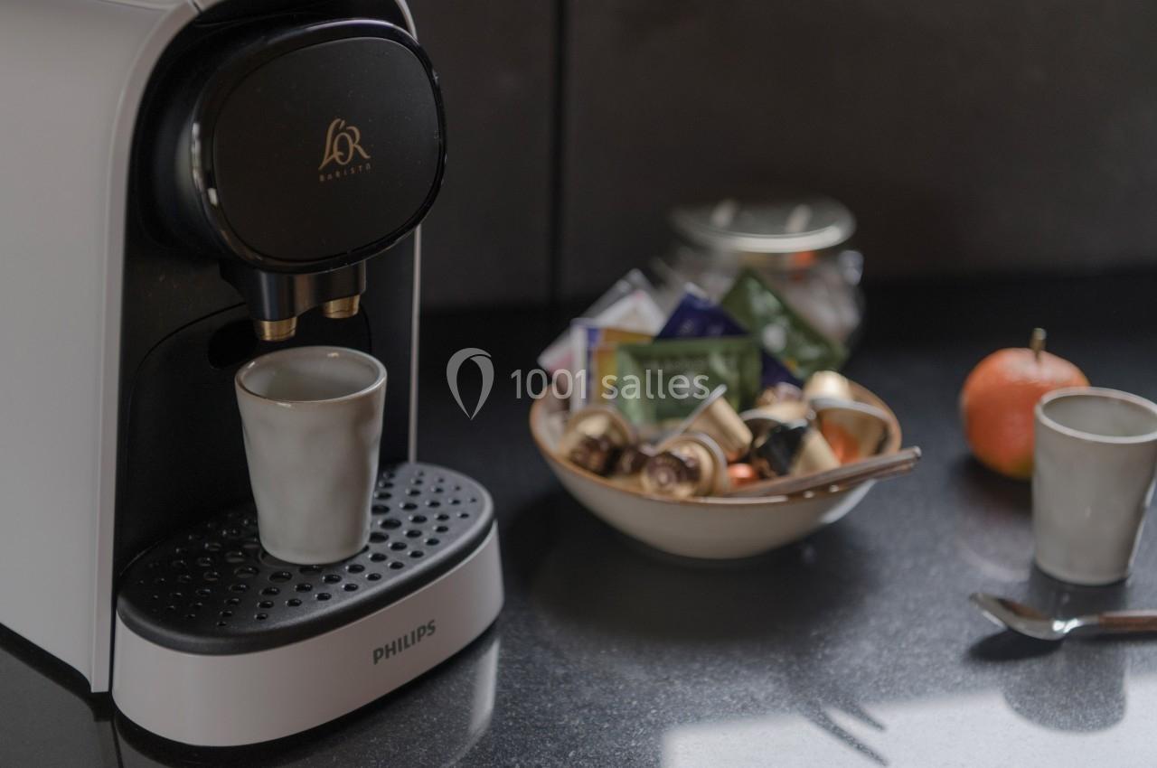 Machine à café avec une tasse en céramique, bol contenant des capsules et sachets de thé sur un plan de travail sombre.