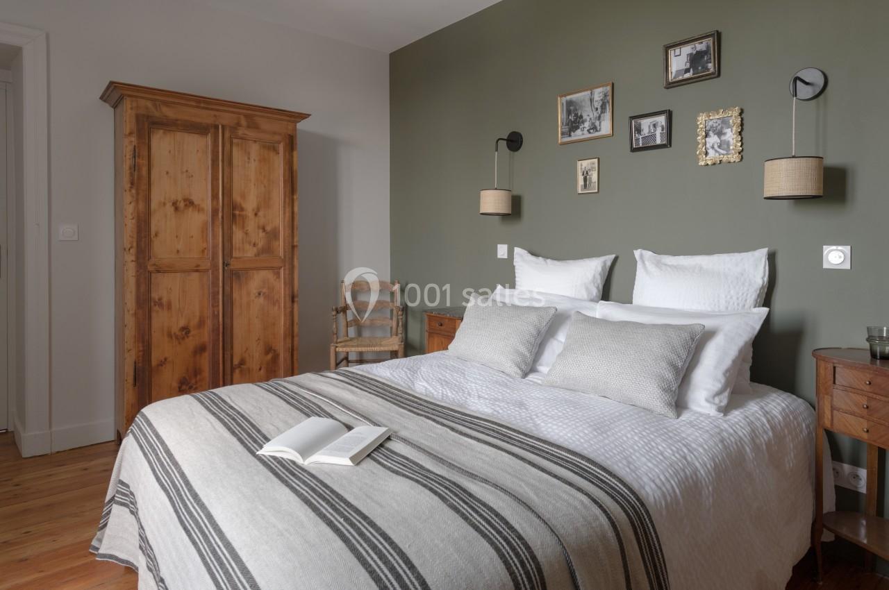 Chambre avec un lit double, linge blanc et coussins, armoire en bois, murs verts décorés de cadres et lampes murales.