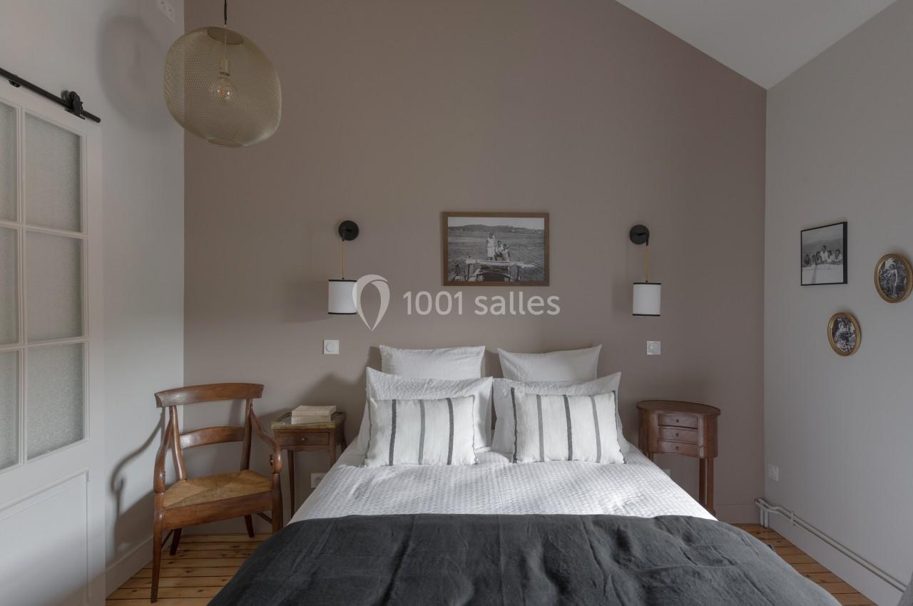 Chambre avec lit double, murs beiges, linge de lit blanc, suspension en rotin, et mobilier en bois.