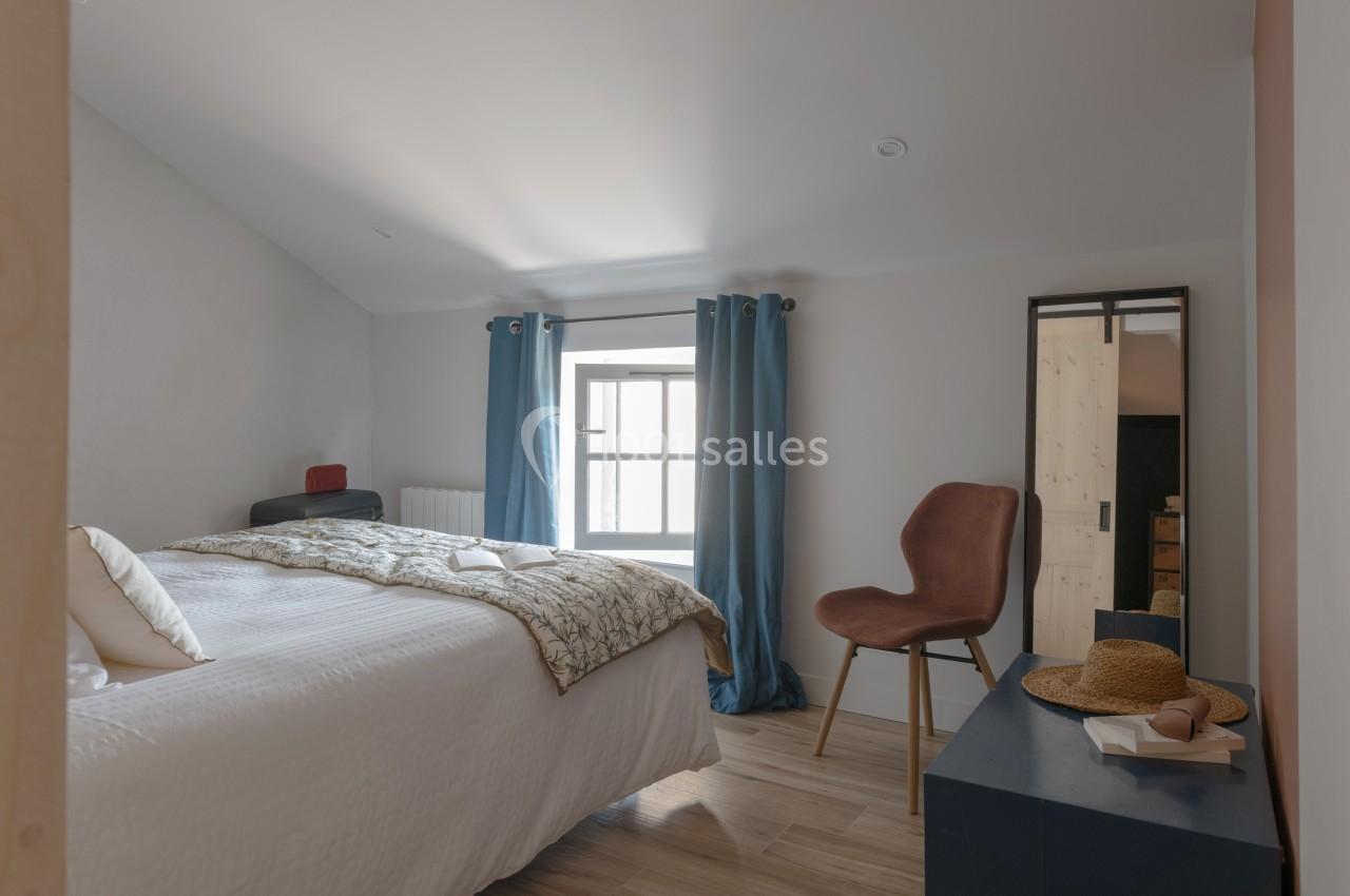 Chambre lumineuse avec un lit double, une chaise, un miroir, des rideaux bleus et un chapeau posé sur une commode.