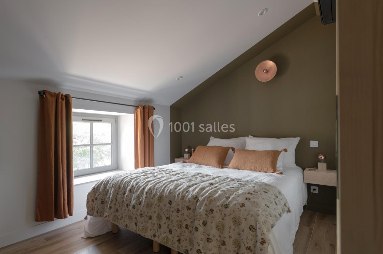Chambre lumineuse avec lit double, mur vert olive, fenêtre avec rideaux orange et parquet au sol.