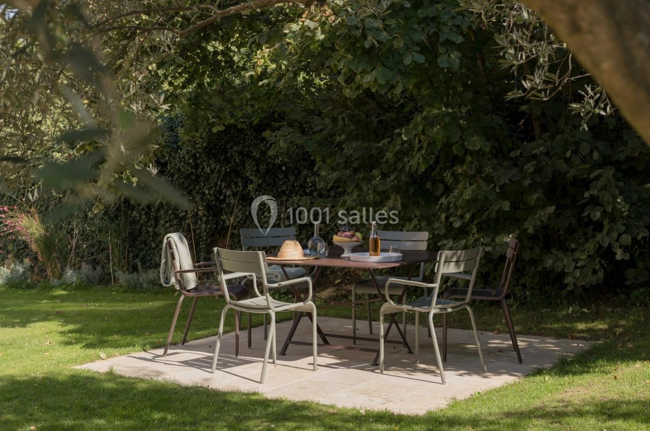 Table de jardin entourée de chaises sur une terrasse en pierre, sous des arbres, avec des plats et boissons disposés.