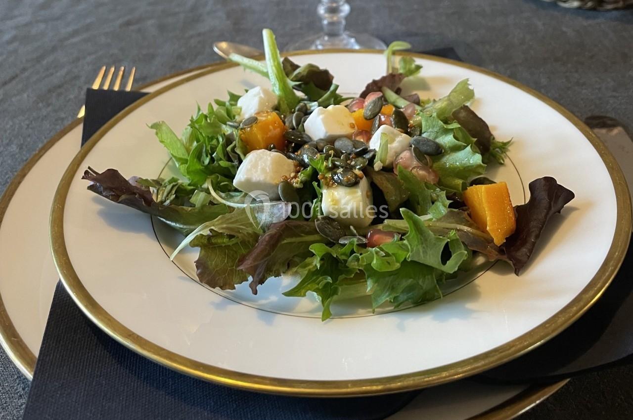 Assiette de salade composée avec feuilles vertes, cubes de fromage, morceaux de potiron et graines, sur une table dressée.