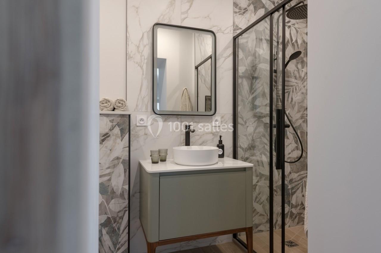 Salle de bain moderne avec vasque blanche, miroir rectangulaire, douche vitrée et carrelage à motifs végétaux.
