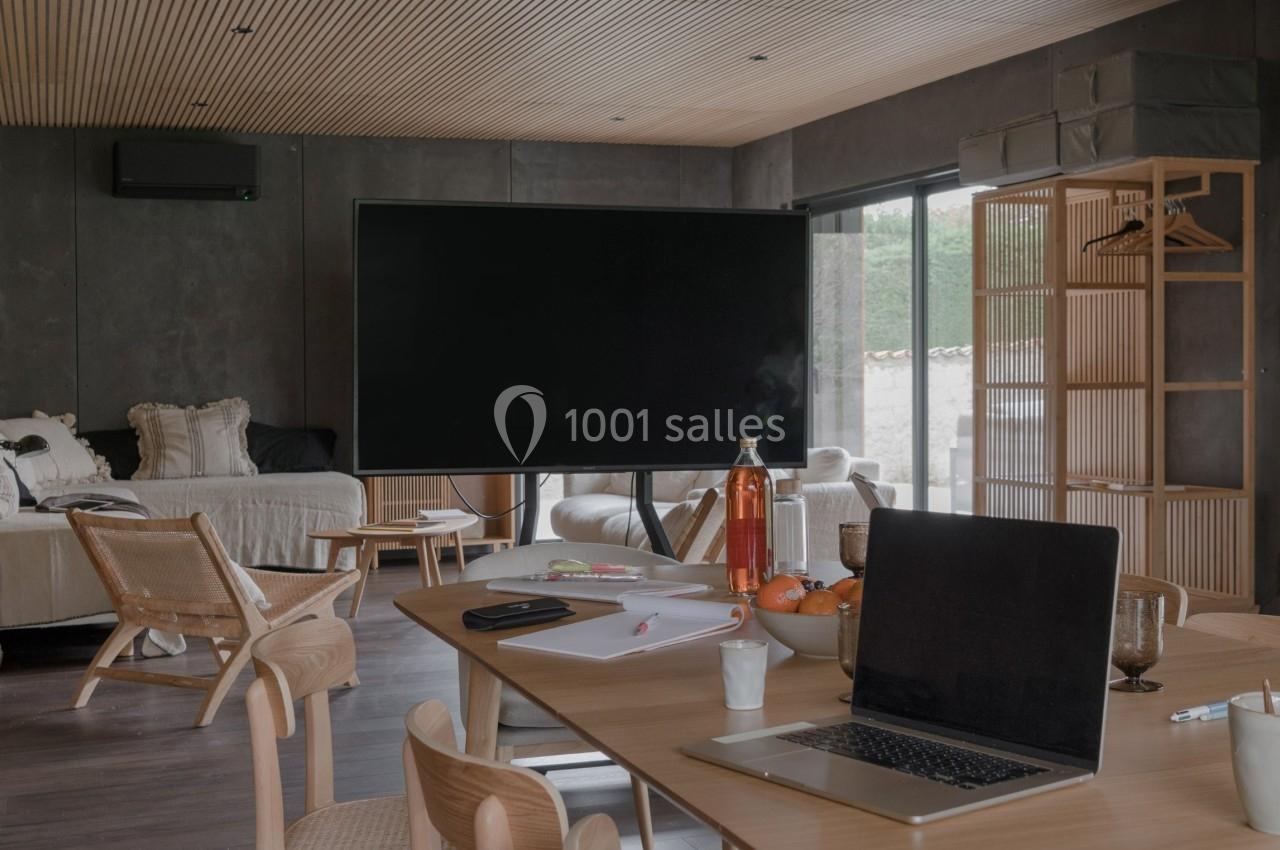 Salle de réunion moderne avec table en bois, chaises, ordinateur portable et écran noir, éclairée par une lumière naturelle.