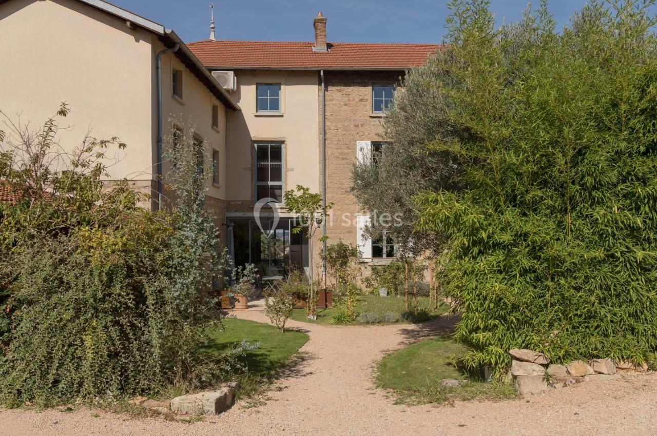 Façade d'une maison en pierre et crépi beige, entourée de végétation et d'un chemin en gravier.