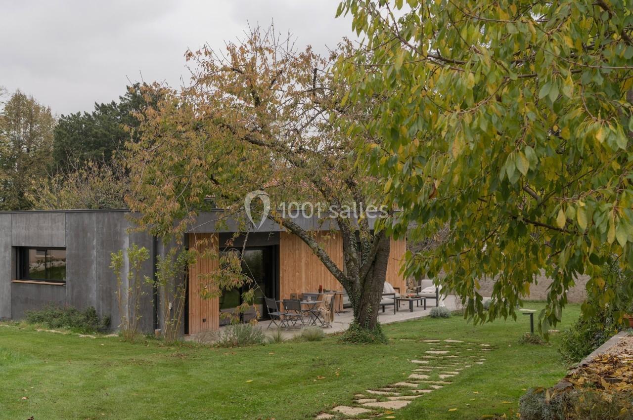 Maison moderne en bois et béton entourée d'un jardin verdoyant avec un arbre au premier plan.