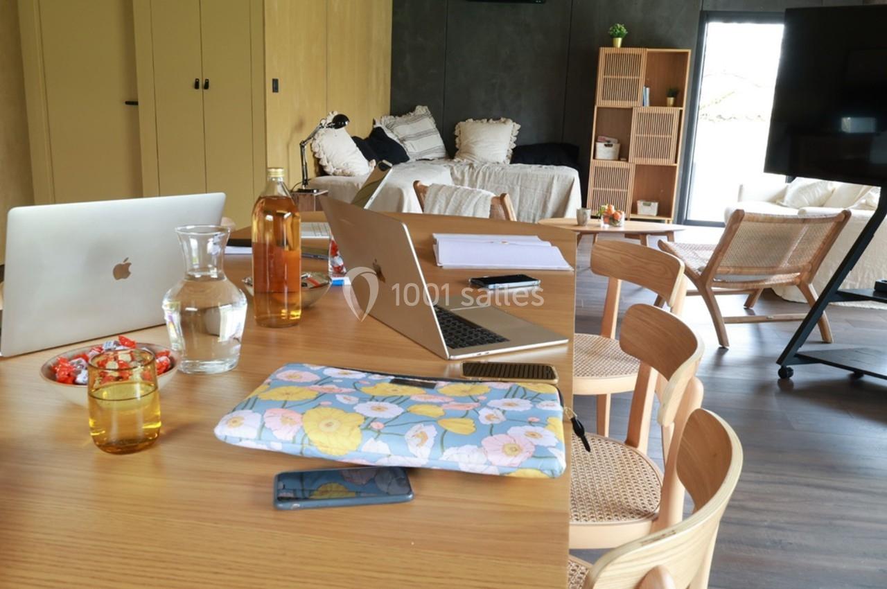 Table en bois avec ordinateurs portables, verre d'eau, bouteille, cahiers et fauteuils dans un salon lumineux.