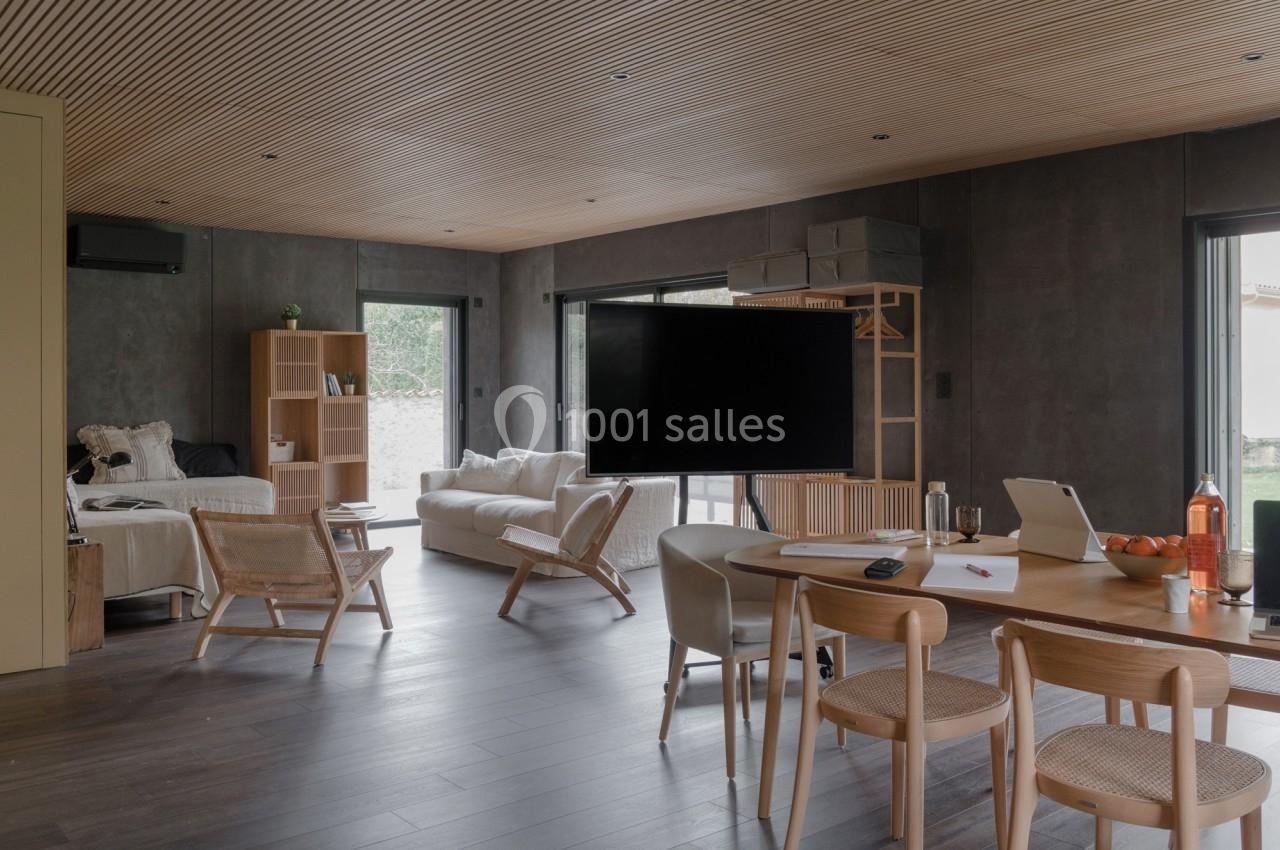 Espace de vie moderne avec mobilier en bois clair, canapé, table de travail et grande télévision sur pied.