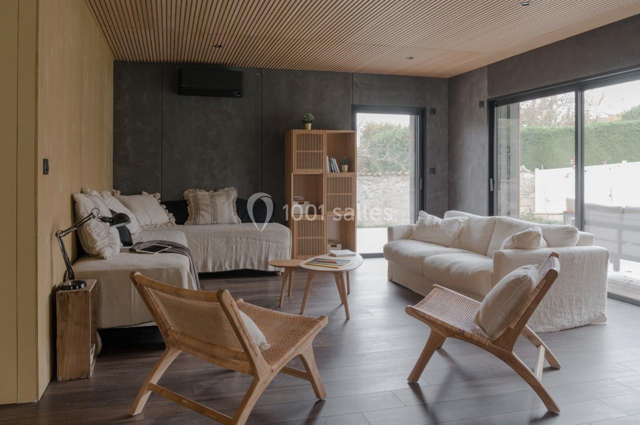 Salon moderne avec parquet foncé, canapé blanc, fauteuils en bois, étagère et grandes baies vitrées donnant sur un jardin.