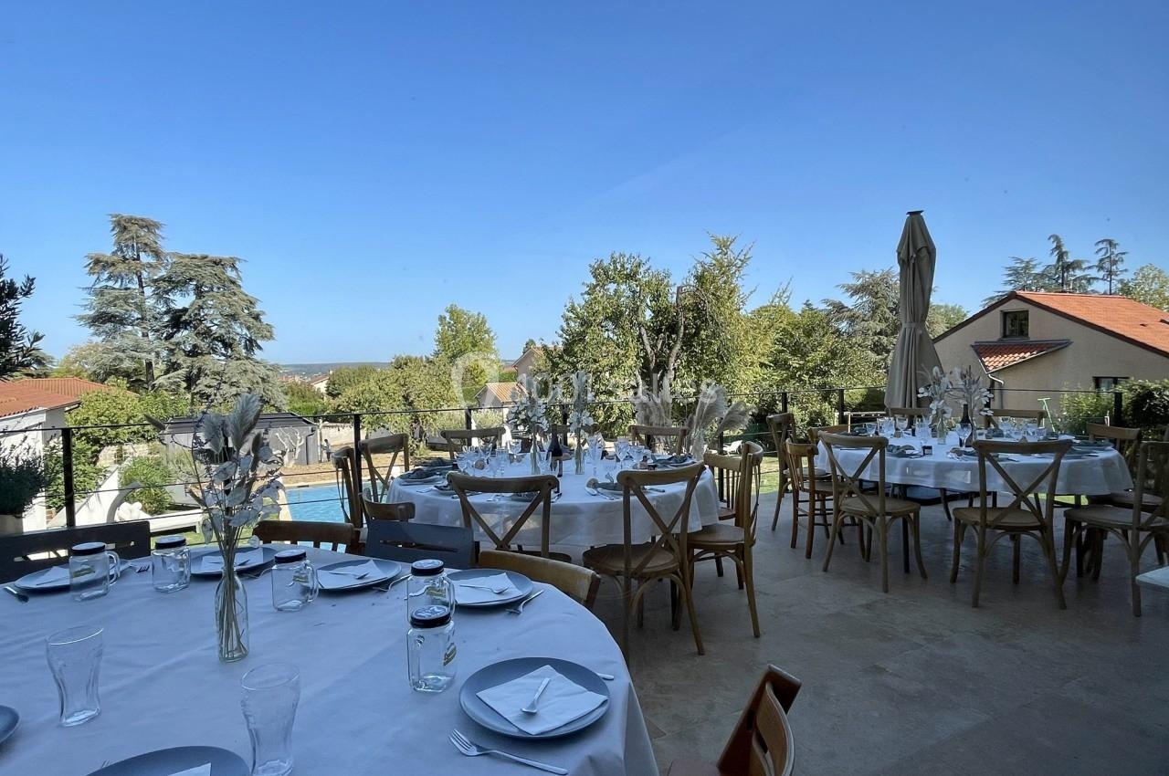 Tables rondes dressées en extérieur avec nappes blanches, verres et décorations, sur une terrasse avec vue sur jardin.
