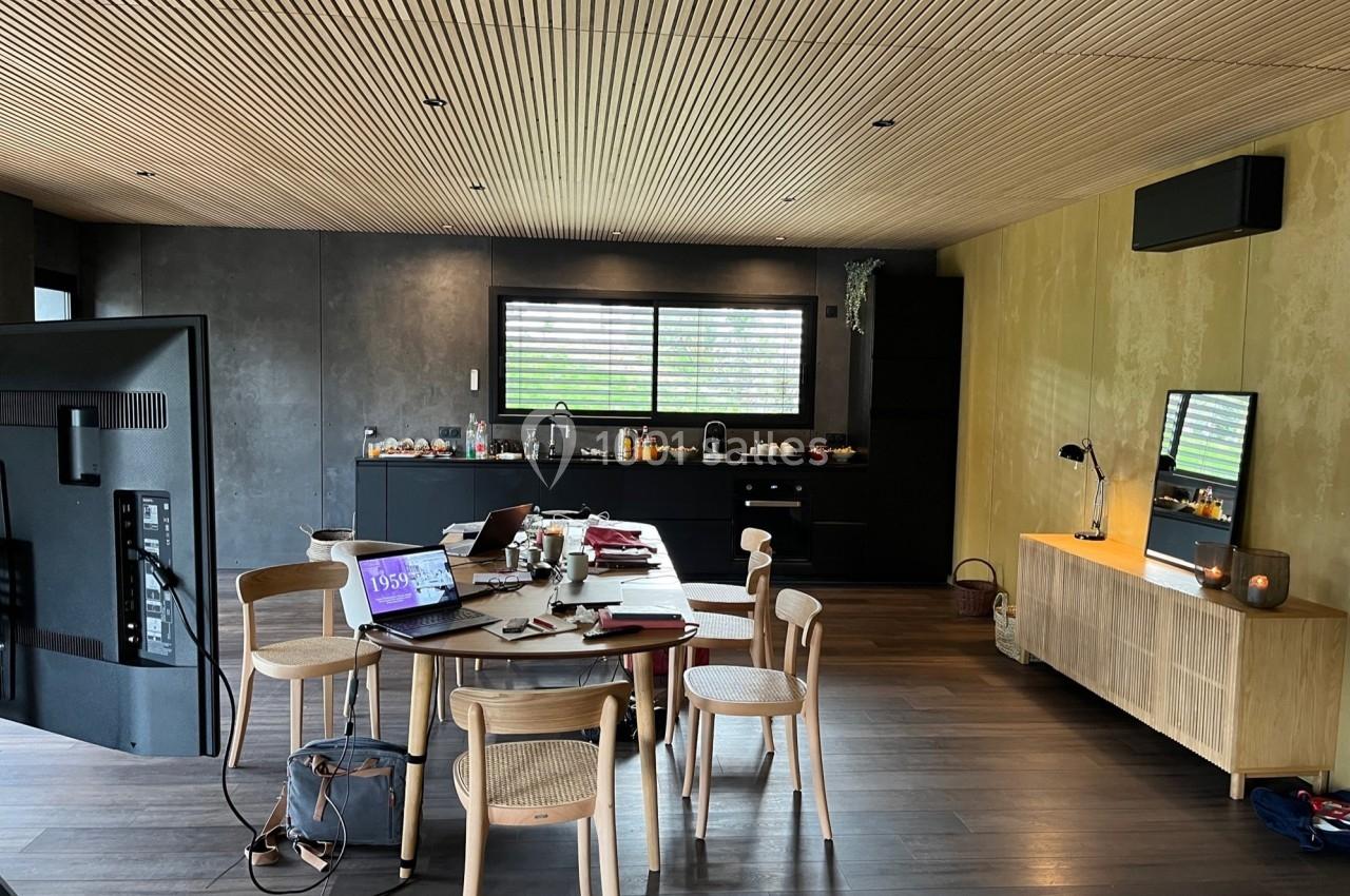 Salle à manger moderne avec table en bois, chaises, cuisine ouverte en arrière-plan et lumière naturelle.