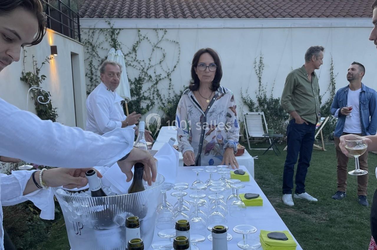 Des personnes discutent et servent des boissons autour d'une table dressée en extérieur, devant un mur blanc.
