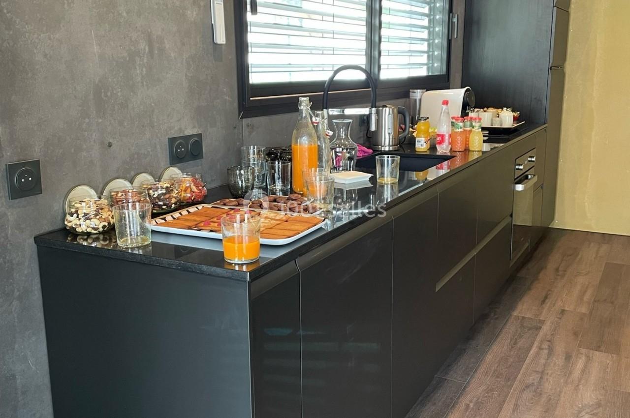 Buffet sur un plan de travail noir avec boissons, viennoiseries et snacks, dans une cuisine moderne éclairée par une fenêtre.