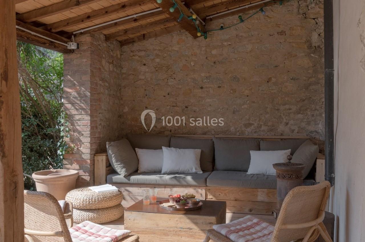 Canapé avec coussins sous un toit en bois, entouré de murs en pierre, avec table basse et fauteuils en osier.