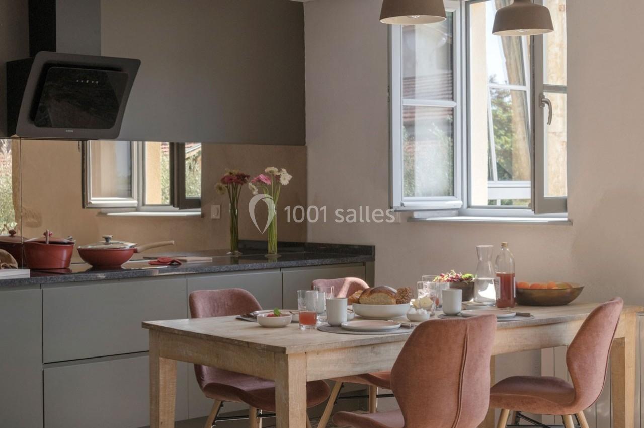 Cuisine lumineuse avec table en bois dressée pour le petit-déjeuner, chaises roses et fenêtre ouverte sur l'extérieur.