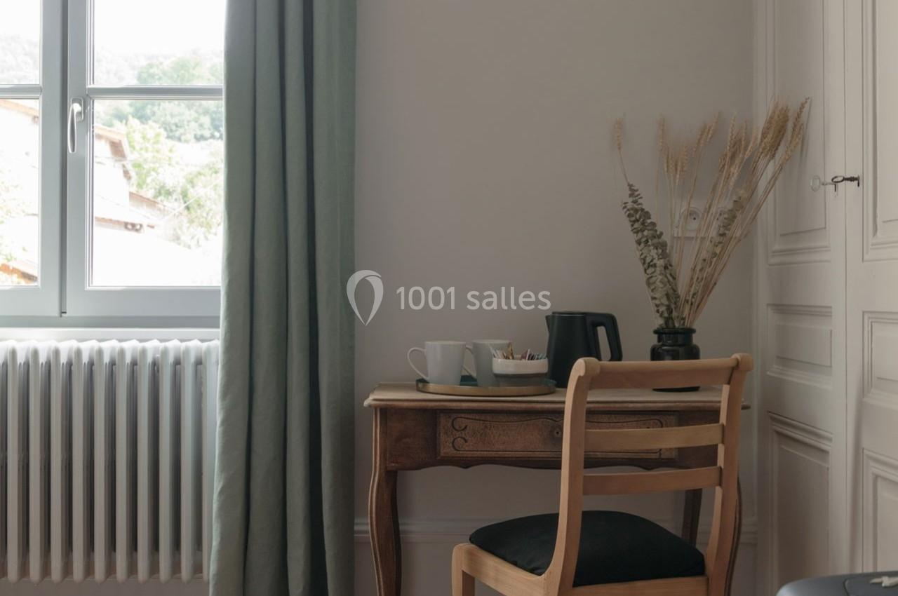 Bureau en bois avec chaise, tasse, bouilloire et vase de fleurs séchées près d'une fenêtre avec rideaux verts.