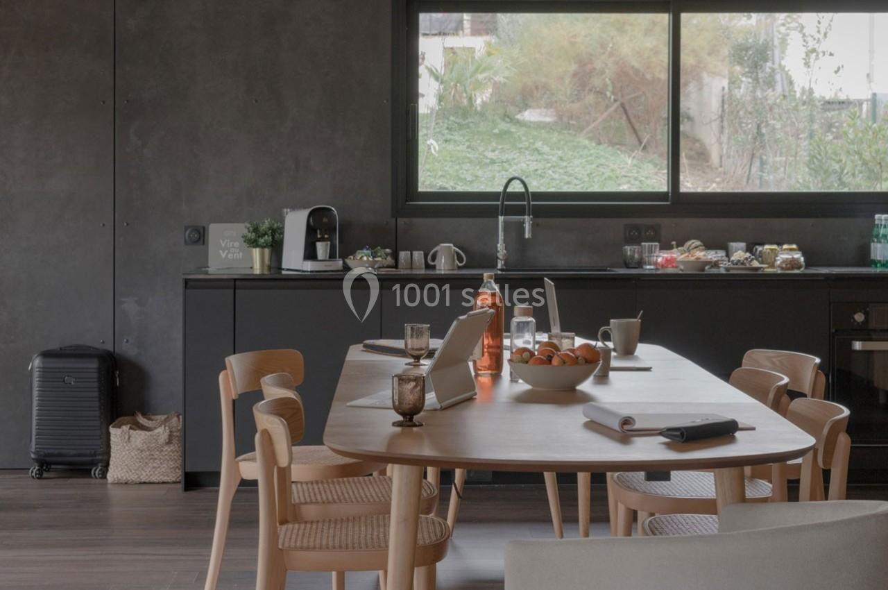 Cuisine moderne avec table en bois dressée pour le petit-déjeuner, vue sur un jardin à travers une grande fenêtre.
