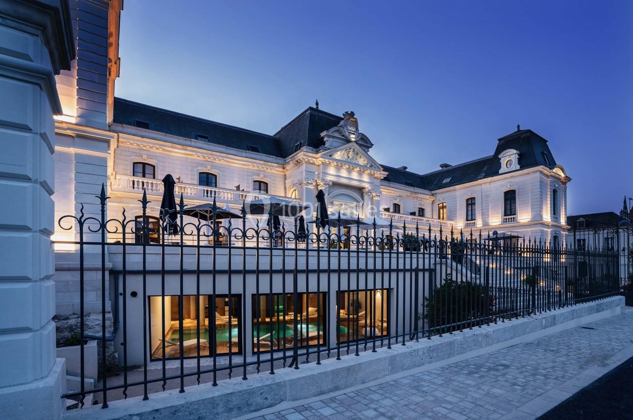 Façade d'un bâtiment historique éclairé en soirée, avec une piscine visible derrière une grille au premier plan.