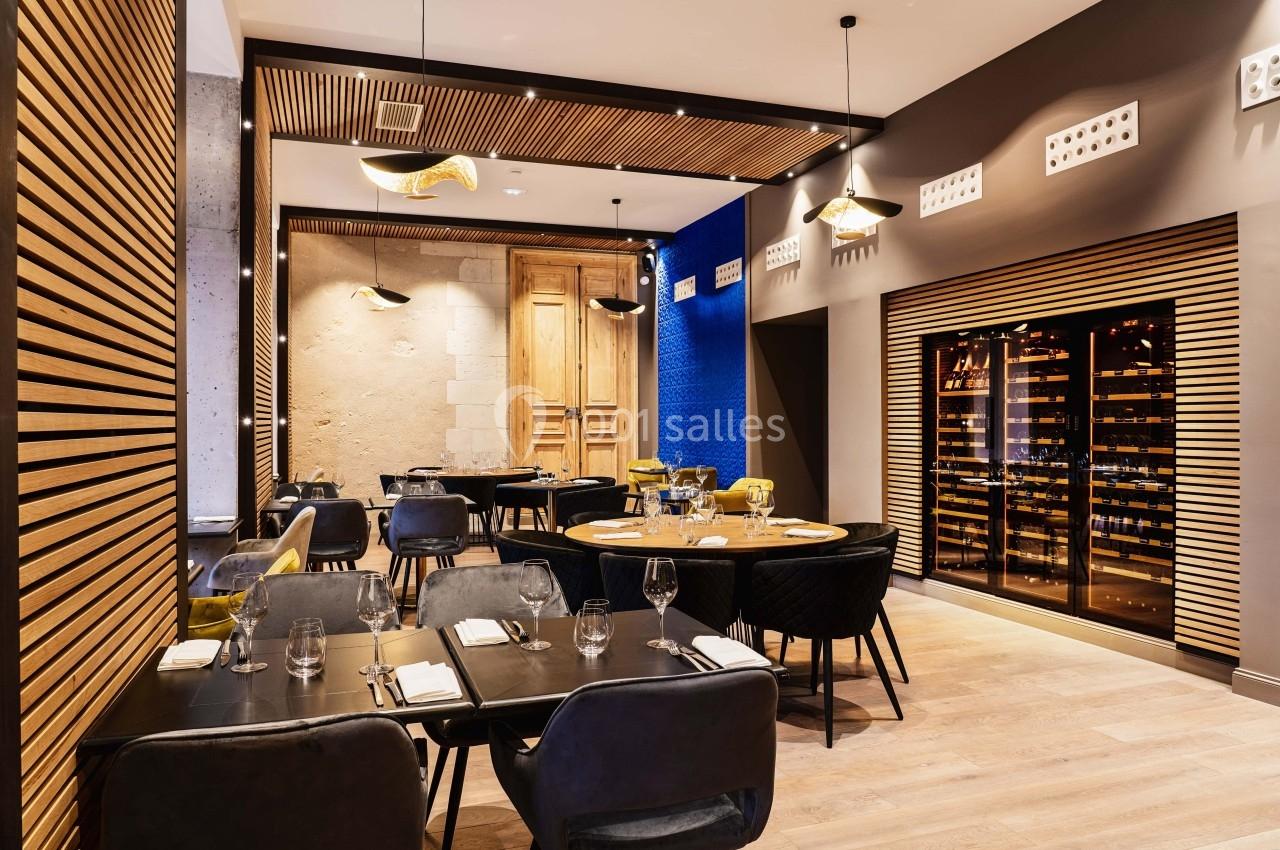 Salle de restaurant moderne avec tables dressées, chaises noires et une cave à vin vitrée sur un mur en bois.