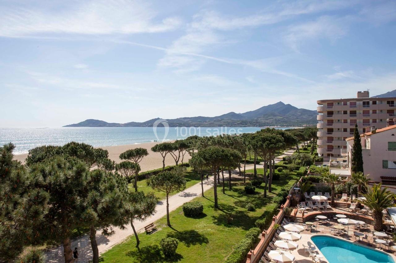 Vue d'une plage bordée d'arbres, avec des montagnes à l'horizon et des bâtiments résidentiels en bord de mer.