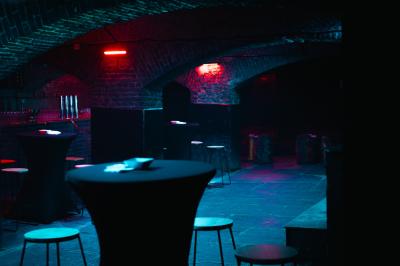 Miniature Location salle Lille (Nord) - La Maison  #6 Salle sombre avec murs en briques, éclairage tamisé, scène en bois et tables basses au premier plan.