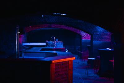 Miniature Location salle Lille (Nord) - La Maison  #8 Salle sombre avec murs en briques, éclairage tamisé, scène en bois et tables basses au premier plan.