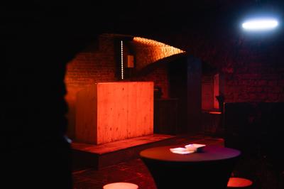Miniature Location salle Lille (Nord) - La Maison  #9 Salle sombre avec murs en briques, éclairage tamisé, scène en bois et tables basses au premier plan.