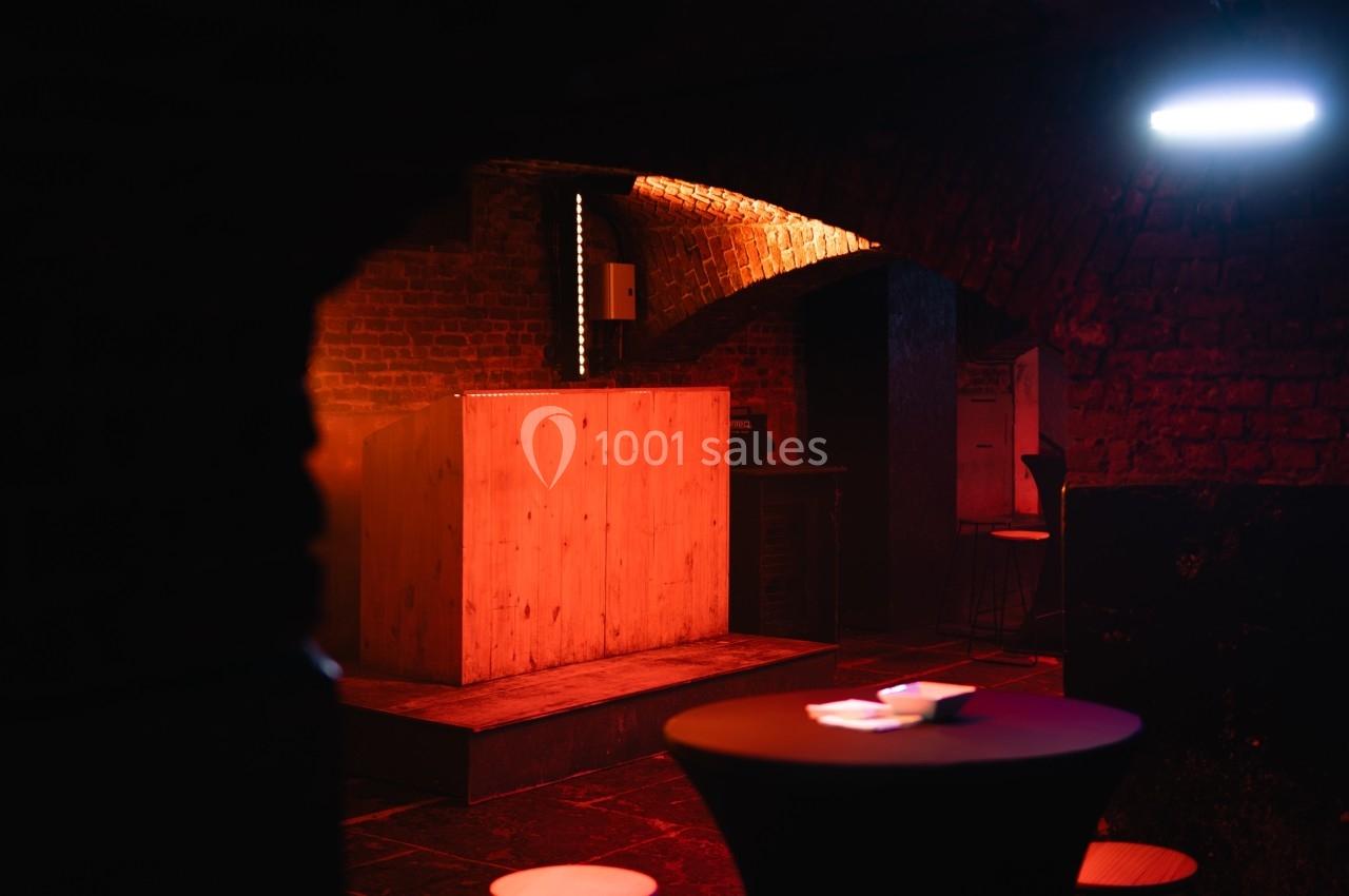 Salle sombre avec murs en briques, éclairage tamisé, scène en bois et tables basses au premier plan.