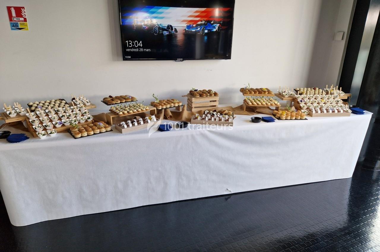 Table garnie de bouchées apéritives variées disposées sur des présentoirs en bois, avec un écran en arrière-plan.