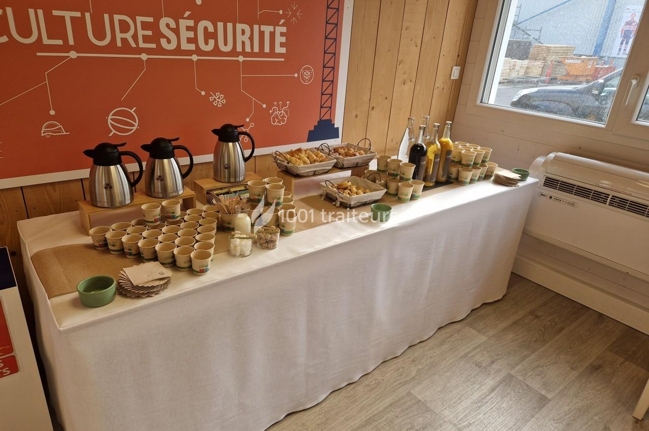 Table dressée avec boissons chaudes, jus, viennoiseries et gobelets dans une salle lumineuse au décor professionnel.