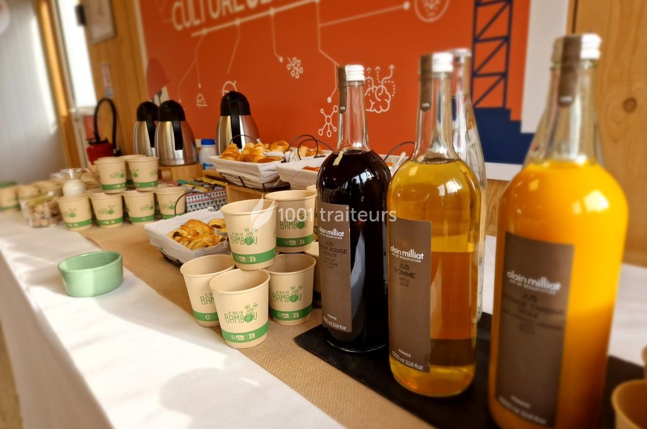 Buffet avec boissons en bouteilles, gobelets en carton, viennoiseries et thermos sur une table blanche.