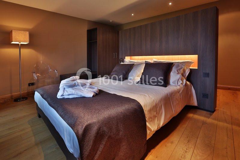 Lit double avec draps blancs et couverture marron, éclairage intégré dans une tête de lit en bois, sol en parquet.