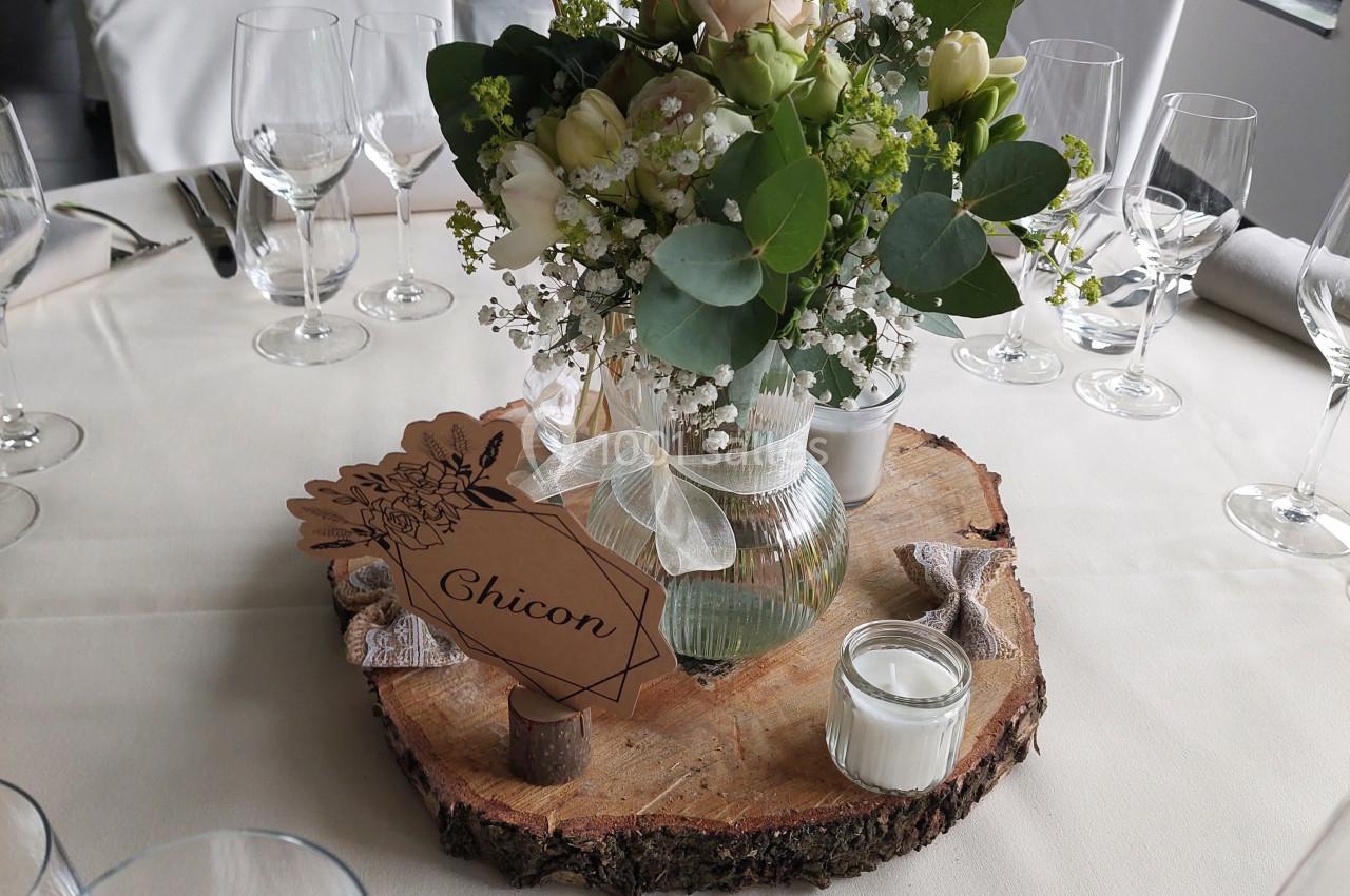 Centre de table avec fleurs blanches et feuillages dans un vase, posé sur une tranche de bois avec une bougie.