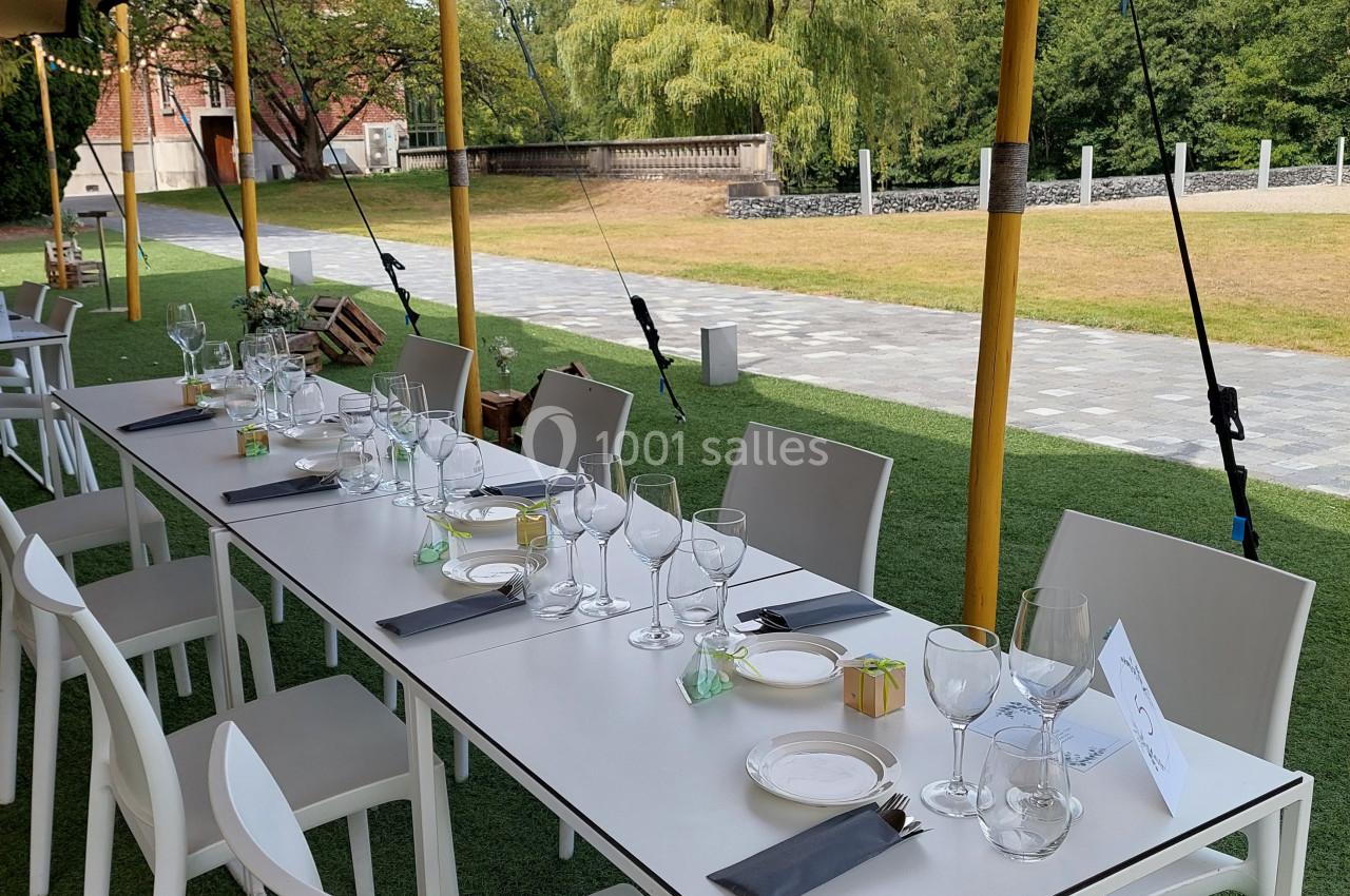 Table dressée en extérieur sous une tente, avec chaises blanches, verres, couverts et serviettes sur une pelouse.