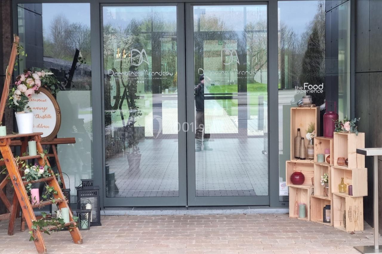 Entrée vitrée d'un bâtiment avec des décorations en bois, fleurs et étagères contenant divers objets.