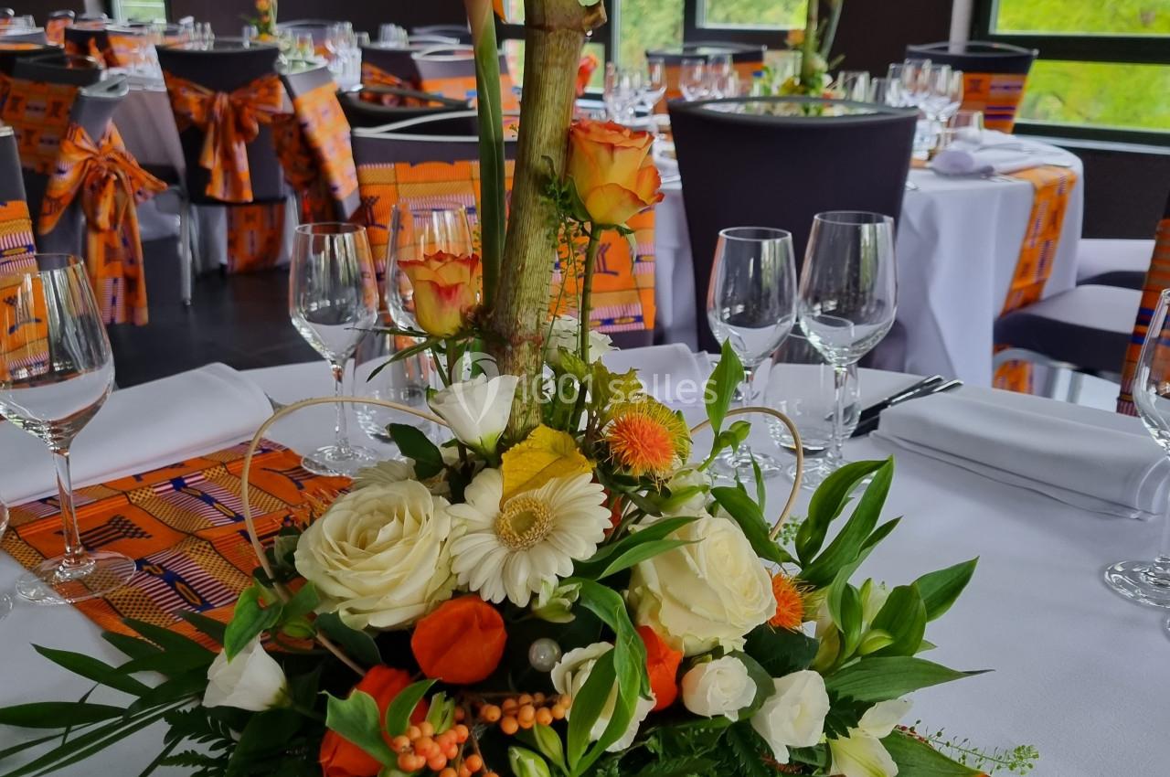 Centre de table floral avec roses, gerberas, feuillage et oiseaux de paradis, dans une salle de réception lumineuse.