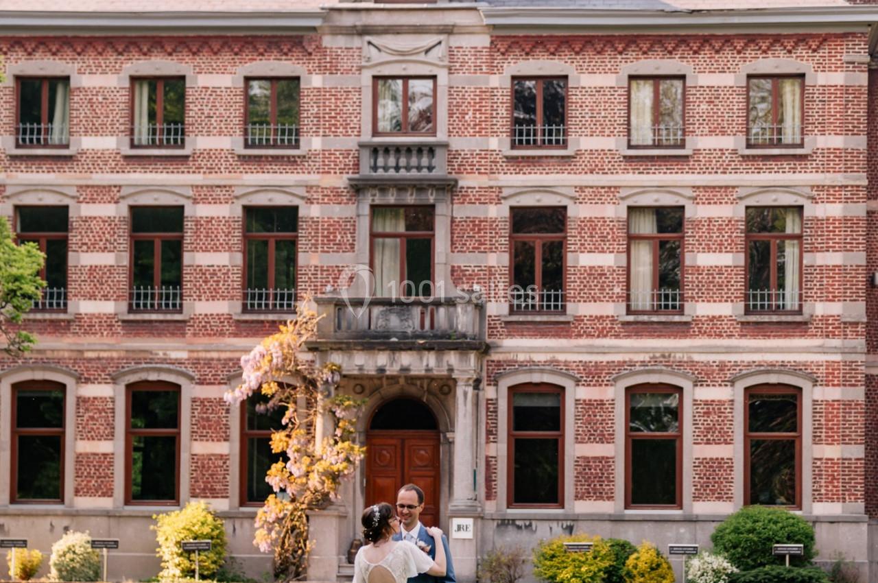 Un couple en tenue de mariage se tient devant un grand bâtiment en briques rouges avec un jardin verdoyant.
