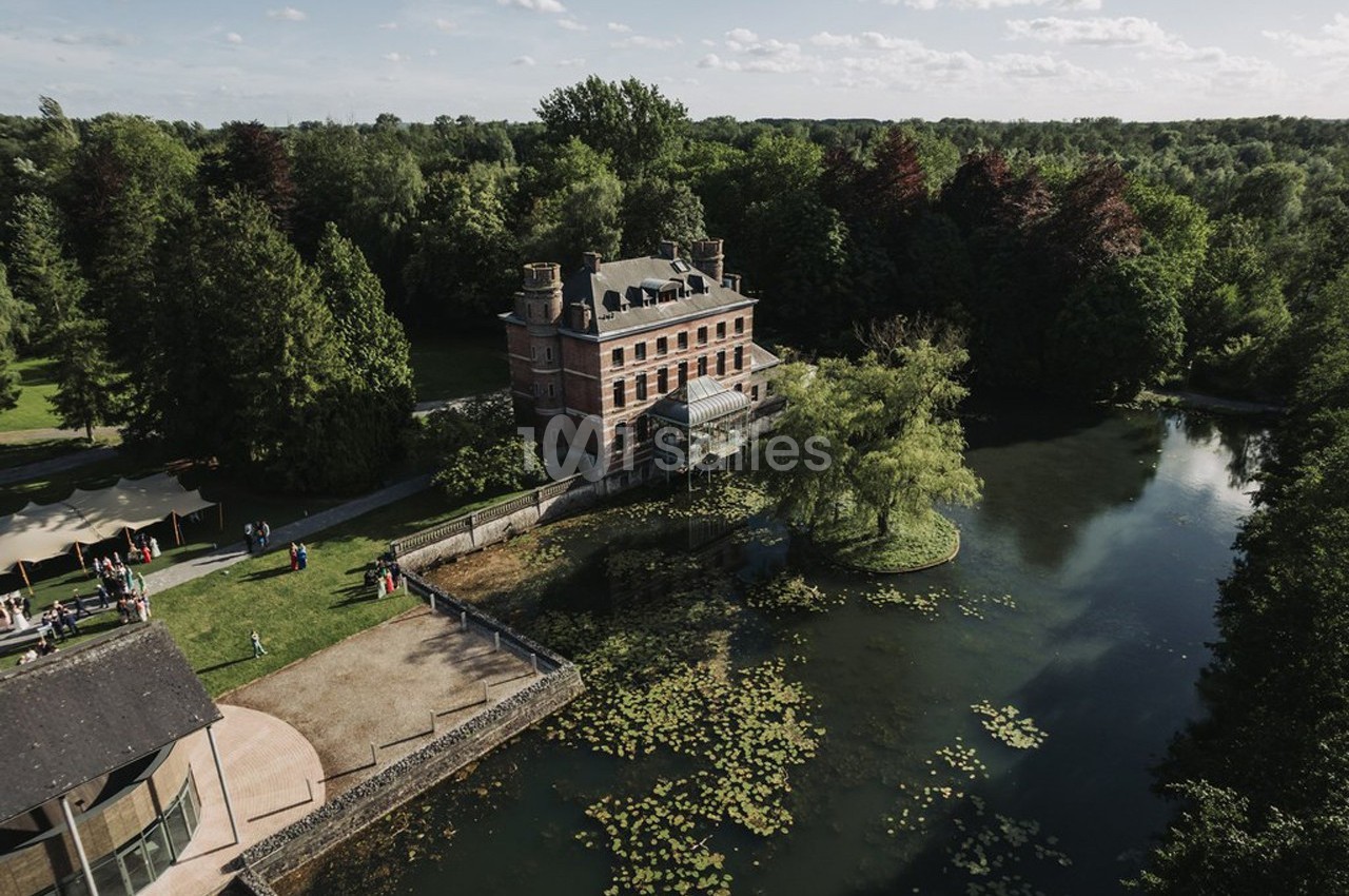 Vue aérienne d'un manoir entouré d'arbres, bordé par un étang avec des nénuphars et une pelouse aménagée.