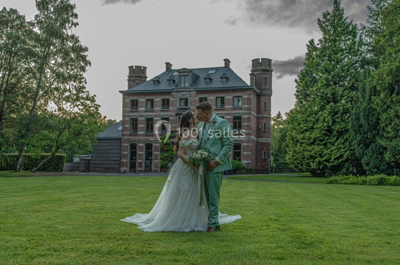 Un couple en tenue de mariage pose devant un château entouré de verdure au crépuscule.