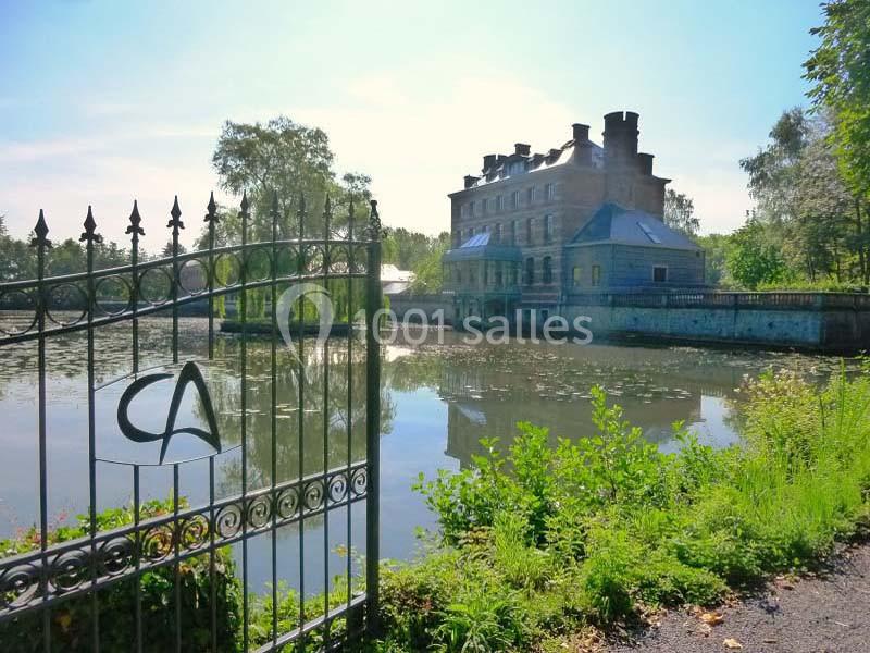 Vue d'un manoir historique en pierre près d'un étang, entouré de verdure et d'une grille en fer forgé.
