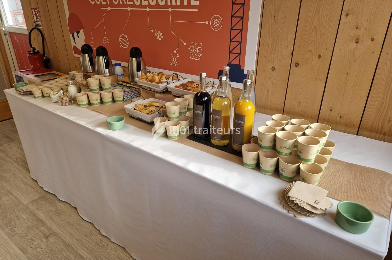 Table avec boissons, viennoiseries et gobelets en carton disposés pour un buffet dans une salle au décor en bois.