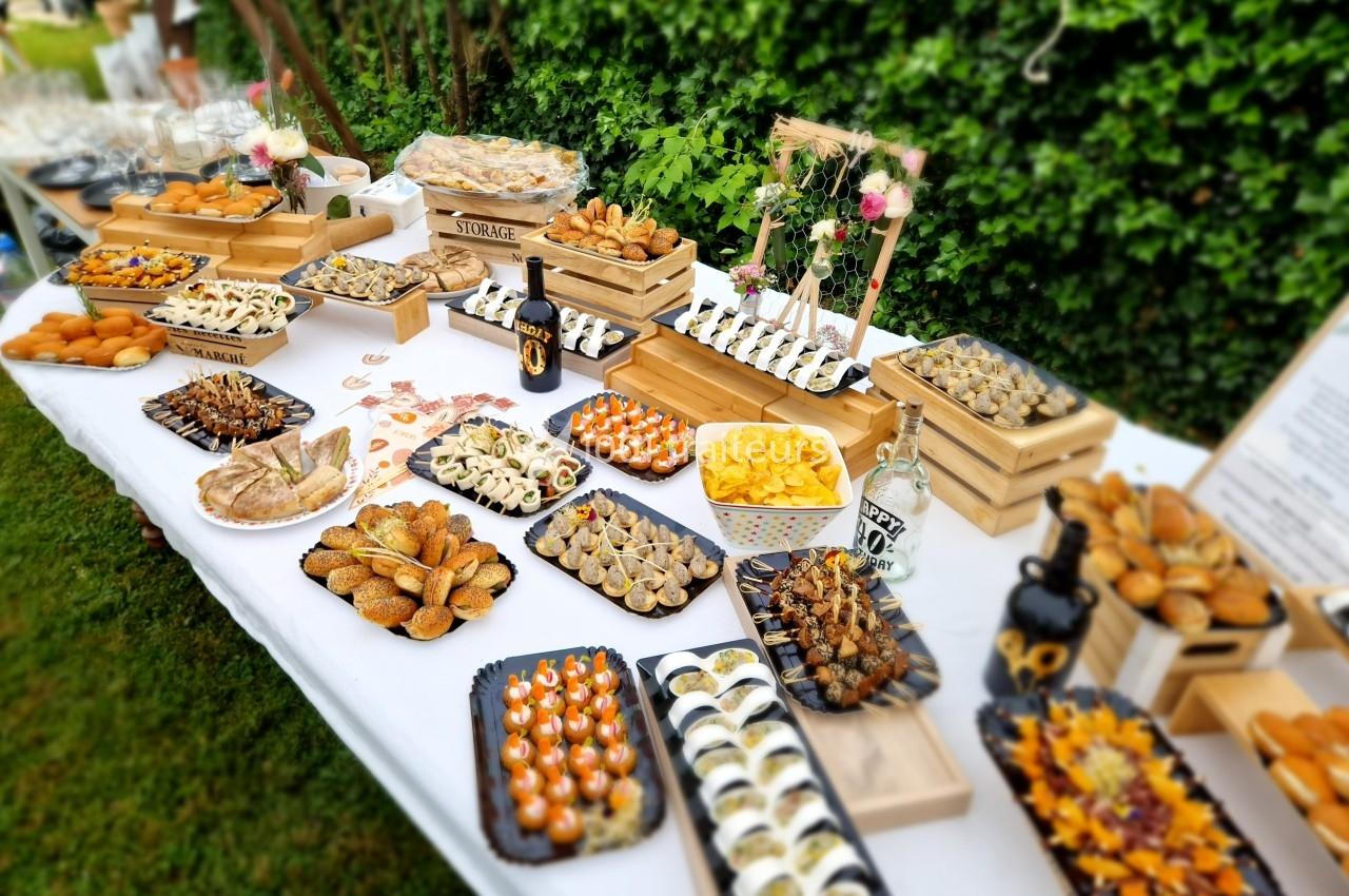 Table garnie de divers amuse-bouches et plats apéritifs disposés sur des plateaux dans un jardin verdoyant.